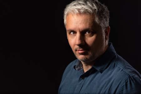 Head and shoulders low key portrait of a bearded middle-aged man looking thoughtfully at the camera over a black studio background with copy spaceの写真素材