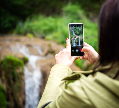 Female hands with red painted nails Taking Photo of a mountain Waterfall with Mobile Phoneの写真素材