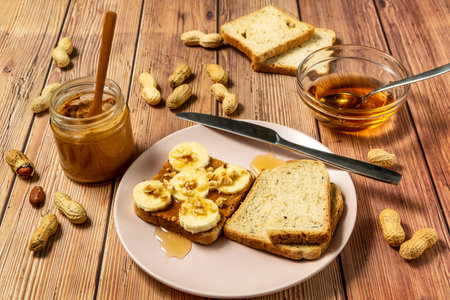 Peanut butter or peanut paste in an open jar and spread on a toast sandwich with banana and honey and peanuts in shells scattered on the table.の写真素材