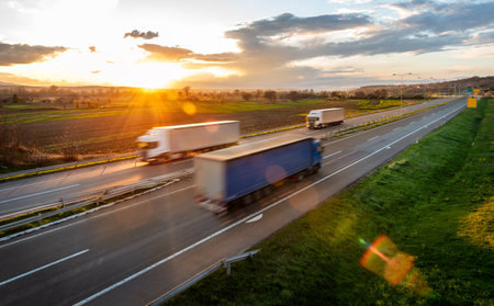 Transportation trucks in high speed driving on a highway through rural landscape at bright sunny sunset. Fast blurred motion drive on the freeway. Freight scene on the motorwayの写真素材