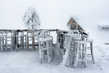 Winter scene in the mountains with snow covered and frozen cottage and outdoor tables and chairs of a restaurant. Frosty outdoor scene of the mountain valley. Beauty of natureの写真素材