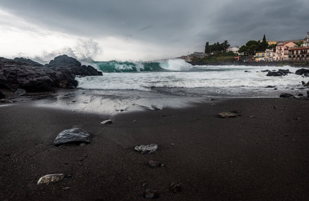 Waves hitting the shore on a cloudy overcast rainy stormy day. Cyclops Coast, black volcanic rocks and send on Sicily, near the town of Acirealeの写真素材