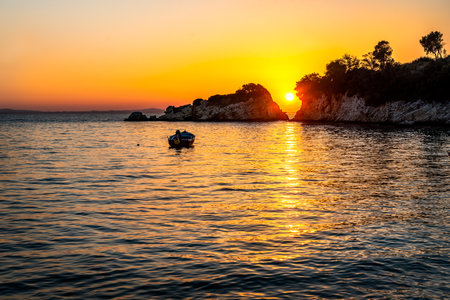 Sunset over the sea. The sun is setting between two rocks in the horizon, with a small motor boat anchored near byの写真素材