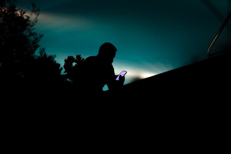 Rear silhouette of a man sitting by the pool at night and looking at his smartphone. Soft focus, silhouette photoの写真素材