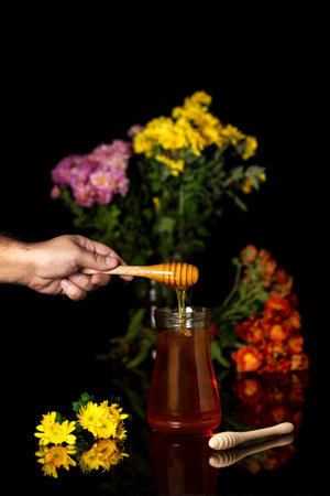 Honey drips from a spoon into a jar on a black background and with wildflowers in the backgroundの写真素材