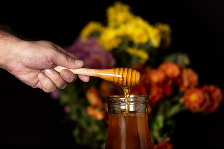 Honey drips from a spoon into a jar on a black background and with wildflowers in the backgroundの写真素材