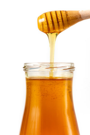 Honey drips from a spoon into a jar on white backgroundの写真素材