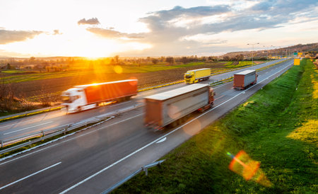 Transportation trucks in high speed driving on a highway through rural landscape, on a beautiful sunset. Fast blurred motion drive on the freewayの写真素材