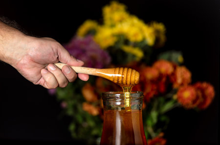 Honey drips from a spoon into a jar on a black background and with wildflowers in the backgroundの写真素材