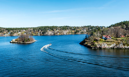 Small Motor boat through in the Stockholm archipelago in Baltic sea with traditional white and red houses at sunny spring evening scenery.の写真素材