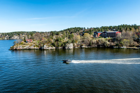 Small Motor boat through in the Stockholm archipelago in Baltic sea with traditional white and red houses at sunny spring evening scenery.の写真素材