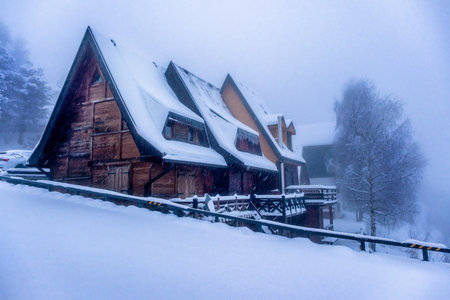 Winter snow falling on wooden cabin in the mountain forest. Winter snow storm blizzard in the mountains. Pine trees covered in snow.の写真素材