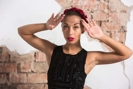 A sexy young girl in a long black dress with heels, with red lips and an attractive smile, a floral rim in her hair poses in the loft of the studio holdind hands near headの写真素材