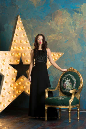 A sexy girl with red lips and curly hair in a luxurious black dress and high heels poses in a rich studio interior with gold and a light starの写真素材