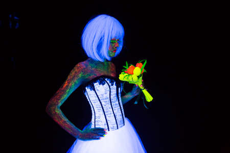 concept of wedding photosession in neon lighting in vogue style bride with bouquet glows in the dark young girl with hairstyle quads and bouquet posing in white wedding dress under the UV rays on a dark background in the studioの写真素材