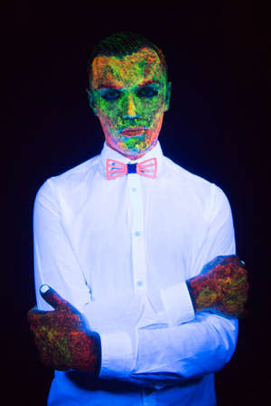 concept of wedding photosession in neon lighting in vogue style groom young guy glows in the dark posing in white wedding suit with a bow tie under the UV rays on a dark background in the studioの写真素材