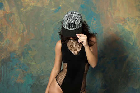 Sexy slender young girl with a sporty body and an attractive breast with curly hair and red lips, with a cap and glasses in a black bodysuit in high heels with bare legs posing in a studio in loft style holdng the cap in the handの写真素材