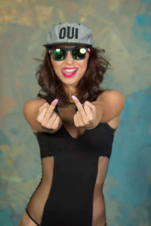 Vogue style Sexy slender young girl showing middle fingers with a sporty body and an attractive breast with curly hair and red lips, with a cap and glasses in a black bodysuit in high heels with bare legs posing in a studio in loft styleの写真素材