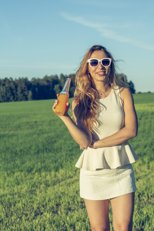 A young slender girl with glasses with curly hair smiles and drinks an alcoholic or non-alcoholic cocktail through a straw from a bottle on a sunny summer day outdoorsの写真素材