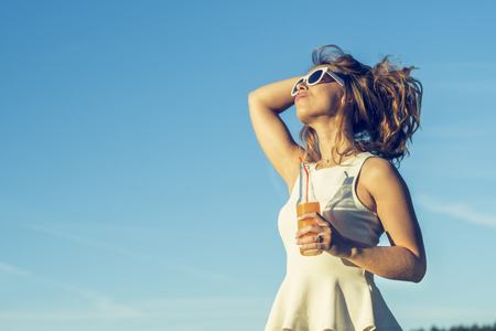 A young slender girl with glasses with curly hair smiles and drinks an alcoholic or non-alcoholic cocktail through a straw from a bottle on a sunny summer day holding her hairの写真素材