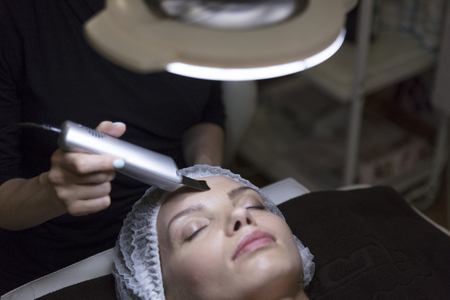 closeup portrait of lovely woman and hand with cosmetic lifting device touching her face under lampの写真素材