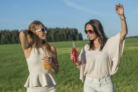 two young slender sisters with glasses andcurly hair smile and drinks an alcoholic or non-alcoholic cocktail through a straw from a bottle on a sunny summer day smilingの写真素材