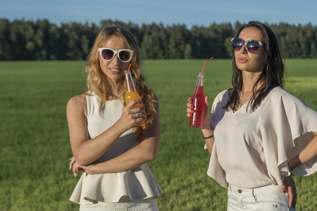 two young slender girls with glasses andcurly hair smile and drinks an alcoholic or non-alcoholic cocktail through a straw from a bottle on a sunny summer day smilingの写真素材