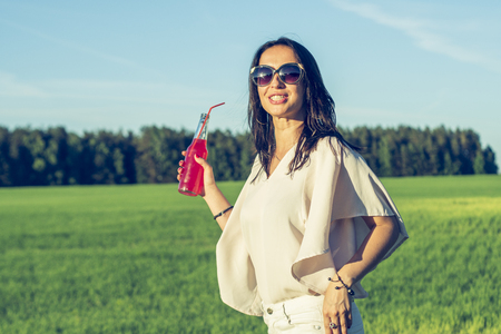 youn slender girl with glasses and curly hair smile and drink an alcoholic or non-alcoholic red cocktail through a straw from a bottle on a sunny summer day smilingの写真素材