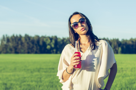 youn slender girl with glasses and curly hair smile and drink an alcoholic or non-alcoholic red cocktail through a straw from a bottle on a sunny summer day smilingの写真素材