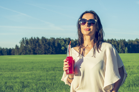 youn slender girl with glasses and curly hair smile and drink an alcoholic or non-alcoholic red cocktail through a straw from a bottle on a sunny summer day smiling outdoorsの写真素材