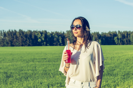youn slender girl with glasses and curly hair smile and drink an alcoholic or non-alcoholic red cocktail through a straw from a bottle on a sunny summer day smilingの写真素材