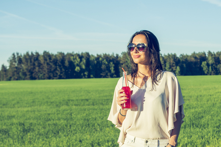 youn slender girl with glasses and curly hair smile and drink an alcoholic or non-alcoholic red cocktail through a straw from a bottle on a sunny summer day smilingの写真素材