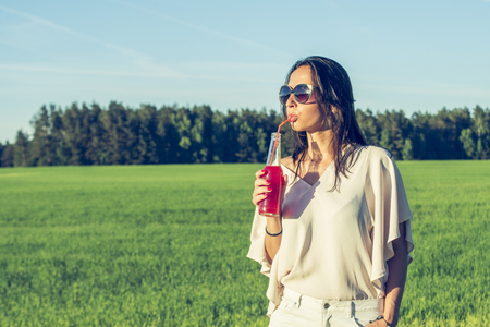 youn slender girl with glasses and curly hair smile and drink an alcoholic or non-alcoholic red cocktail through a straw from a bottle on a sunny summer day smiling on sunsetの写真素材