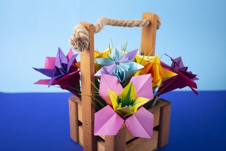 Handmade colored paper flowers origami bouquet paper craft art in a basket with grass in the studio on colored backgroundの写真素材