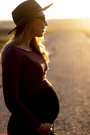 Happy pretty pregnant woman in hat, tight dress smiles in sunlight. Outdoors