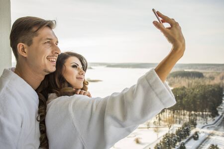 Beautiful sexy girl and handsome man in white bathrobes have fun taking selfiの写真素材
