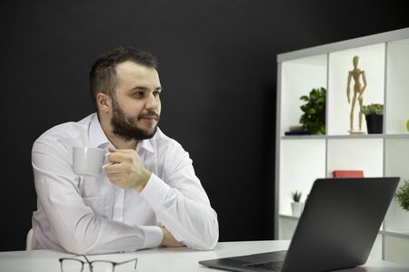 Dreaming handsome man with cup of coffee sitting at table in front of laptop