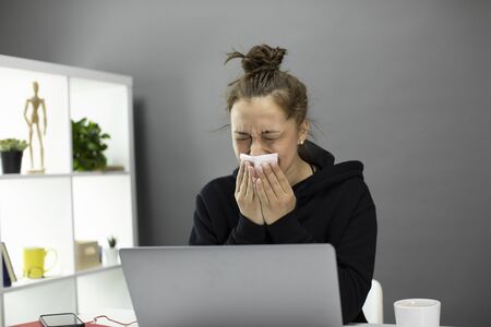 Sick young woman sitting at computer in home office, feels unwell and sneezes