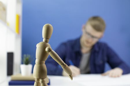 Thoughtful focused young male designer draws with pencil in studio with wooden dummy on blue background. Selective focus on wooden dummy creative process conceptの写真素材