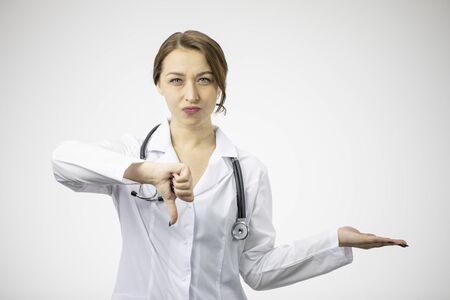 Beautiful female displeased doctor in medical uniform showing dislike sign holding object or product on open palm on white background. Health, healthcare, insurance and medicine recommendation conceptの写真素材