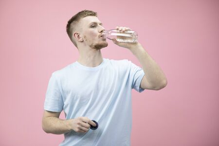 Portrait of young handsome fit body man in t-shirt drinking fresh water from bottle isolated on pink...