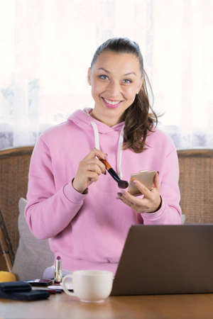 Attractive smiley girl with long dark hair dressed in pink hoodie works remotely with laptop...