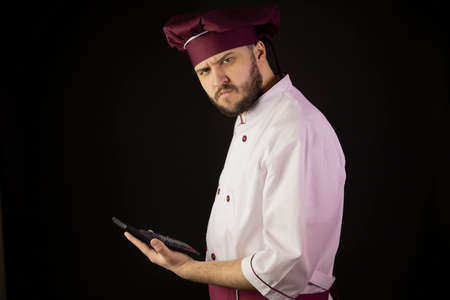 Confused chef man in uniform holds calculator suspiciously looks at camera. Ð¡ook in apron on black background. Restaurant bankruptcy, food industry crisis, business problems, incur loss, breakevenの写真素材