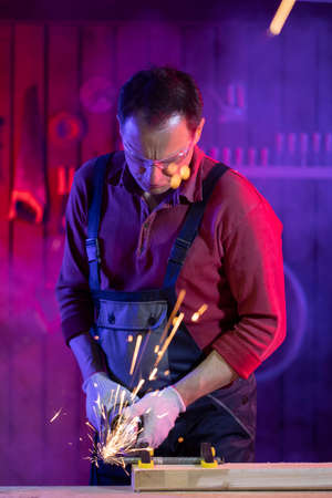 handsome middle-aged male craftsman in jumpsuit protective goggles and gloves grinding metal with...