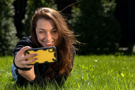Attractive brunette model with long hair dressed in black hoodie lying on green lawn makes selfies...