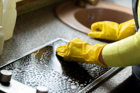 Hands in yellow gloves wipe surface of black induction stove close up. House cleaning concept. Household chemicals concept. Removal of contaminants with chemical solventsの写真素材