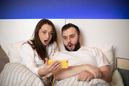Shocked and scared millennial couple using smartphone lying on bed together, surprised boyfriend and...