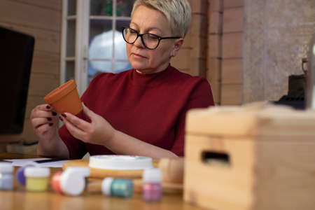 attractive middle-aged woman paints flower pot in cozy country house table view. Concentrated woman...