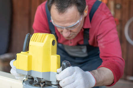 Handsome focused on craftsman processes on wooden workpiece with milling tool close up at cottage workshop. Processing of Wooden beam with milling woodworking machine. Working with hand tool on woodの写真素材