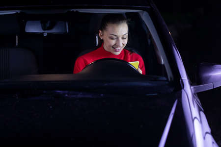 smiling brunette long hair girl in car driving at night smiling reading messages holding smartphone shares photos in social networks, uses free internet connection. safe driving concept. copy spaceの写真素材